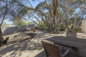 Een terras buiten met een houten tafel en stoelen, omgeven door bomen en een grindpad.