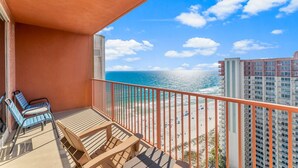 Un balcón con vista al océano y a la playa, con una mesa y sillas de madera, y una barandilla roja.