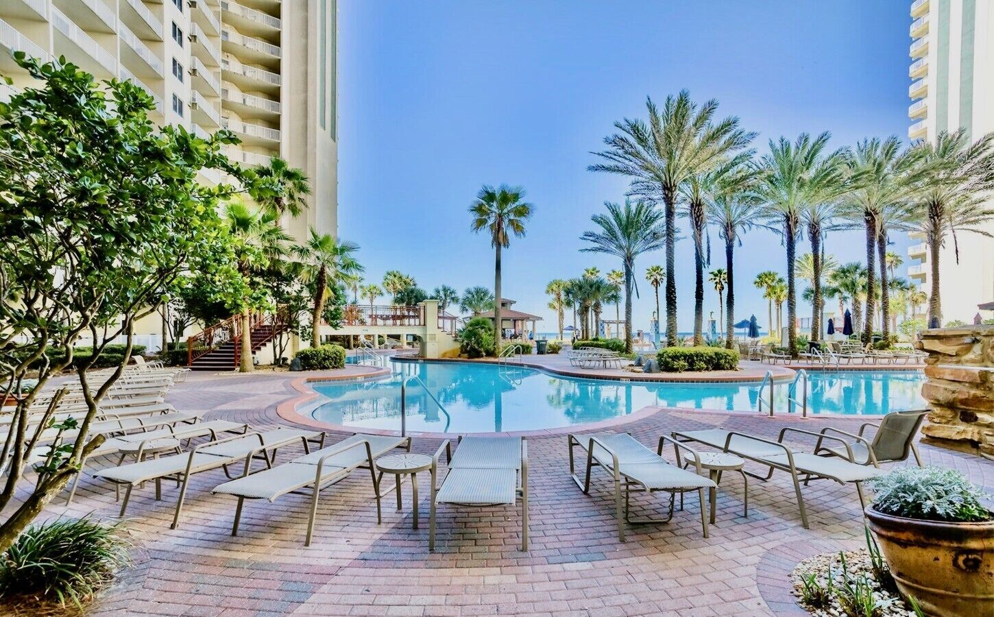 A hotel pool area with lounge chairs, palm trees, and a clear blue sky.