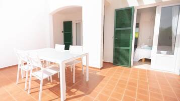 Una mesa blanca con sillas en un patio con suelo de baldosas, contraventanas verdes y un armario blanco.