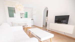 Un salon moderne avec un canapé blanc, une table basse blanche, un téléviseur à écran plat posé sur un meuble blanc, et un coin repas avec une table et des chaises blanches.