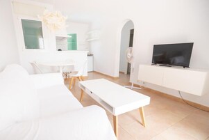 Una sala moderna con un sofá blanco, una mesita de centro blanca, un televisor de pantalla plana sobre un mueble blanco y una zona de comedor con una mesa y sillas blancas.