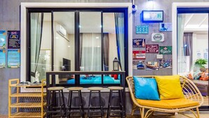 Ein Zimmer mit großem Fenster, einem Essbereich mit Stühlen und Tisch, einem Korbssofa mit Kissen sowie Wanddekorationen, darunter Bierlogos.