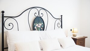 Una cama bien tendida con sábanas blancas y almohadones decorativos, un cabecero metálico con una obra de arte floral, y una mesita de noche con una lámpara.