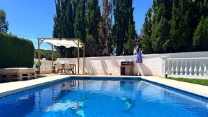 En swimmingpool med et pergolaområde og siddepladser, omgivet af et hvidt hegn og høje træer.