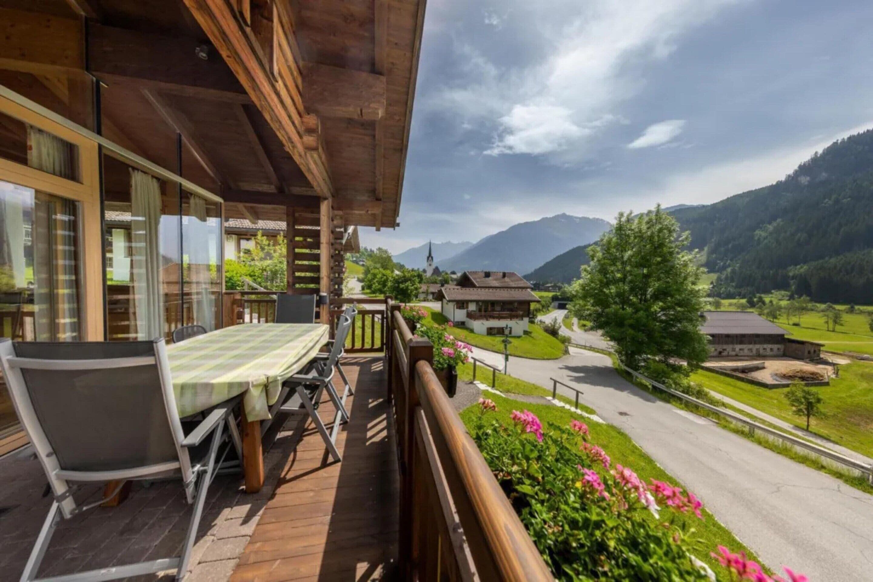 Un balcón de madera con una mesa y sillas, con vistas a un paisaje rural con casas y montañas.