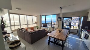 Een ruime woonkamer met een grote bank, een houten eettafel en uitzicht op de stad en zee door grote ramen.