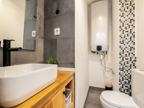 Ein modernes Badezimmer mit einem großen weißen Waschbecken, einem Holz-Waschtisch, einer Toilette und einer Duschkabine mit Wandfliesen aus Mosaik.