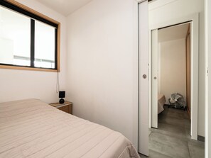 Ein Schlafzimmer mit einem Bett, einem Nachttisch mit Lampe, einem Fenster und einer Tür zu einem Flur.