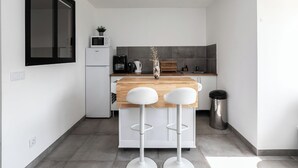 Eine moderne Küche mit einem weißen Kühlschrank, einer Mikrowelle, einer Kaffeemaschine und einem Spülbecken. Zwei weiße Barhocker und eine Holz Arbeitsplatte sind vorhanden.