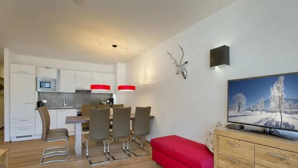 A modern kitchen with white cabinets, a dining area with chairs, a red bench, and a television on a wooden cabinet. - Stylish Neukirchen Home for 8 Guests (Neukirchen am Grossvenediger)