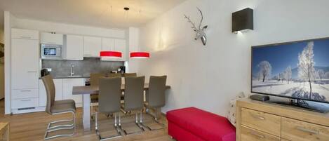 Una cocina moderna con armarios blancos, un comedor con sillas, un banco rojo y un televisor sobre un mueble de madera.