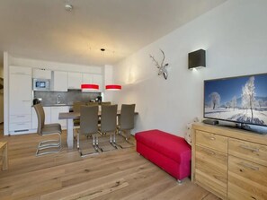 Une cuisine moderne avec des meubles blancs, un coin repas avec des chaises, un banc rouge et un téléviseur posé sur un meuble en bois.