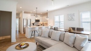 Cocina moderna con isla central, armarios blancos y electrodomésticos de acero inoxidable. Cuenta con un comedor con mesa y sillas de madera, y una sala de estar con un sofá grande y un reposapiés.