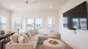 Una sala de estar moderna con un televisor de pantalla plana grande, un sofá color beige, una mesa de comedor y vistas de la ciudad a través de amplias ventanas.