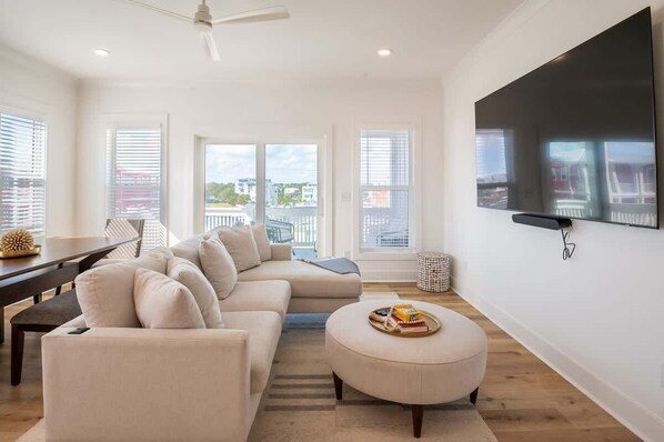 A modern living room with a large flat-screen TV, a beige sofa, a dining table, and a view of the city through large windows.