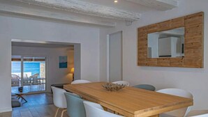Un comedor con mesa de madera y sillas blancas, un espejo grande en la pared y vista al mar a través de una puerta abierta.