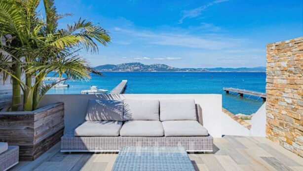 Zona junto a la piscina con sofá, palmera y vista al mar.