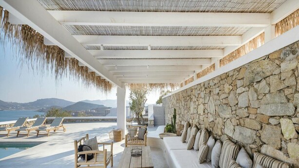 Un espace de détente extérieur, baigné de lumière, avec un mur en pierre, des fauteuils, une table en bois et une vue sur la mer.