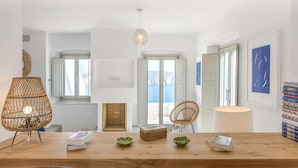 Un intérieur moderne avec une table à manger en bois, des chaises en osier et une vue sur la mer grâce à de grandes fenêtres.
