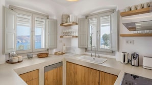 Une cuisine moderne avec des meubles en bois, un lave-vaisselle encastré, un évier et une cuisinière. On y trouve des fenêtres offrant une vue sur la côte, ainsi que des étagères remplies d’ustensiles de cuisine.