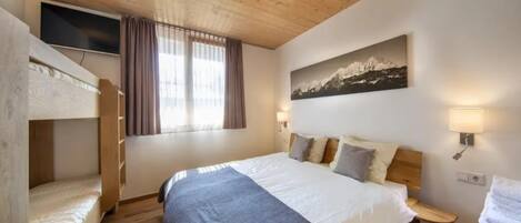 A bedroom with a bunk bed, a large bed with pillows, a mounted TV, a window with curtains, and a painting on the wall.