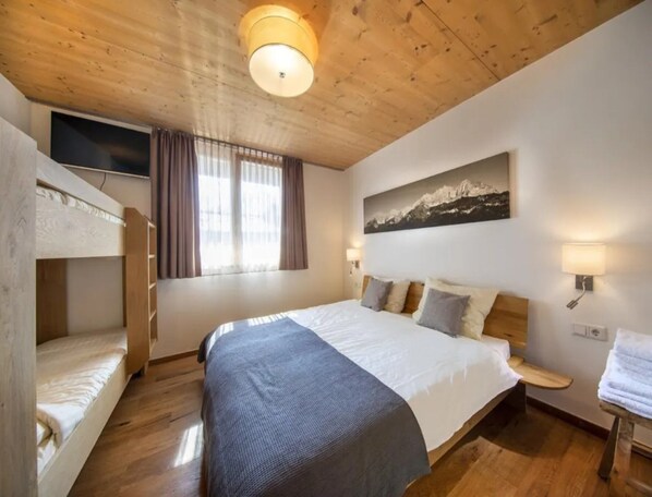 A bedroom with a bunk bed, a large bed with pillows, a mounted TV, a window with curtains, and a painting on the wall.