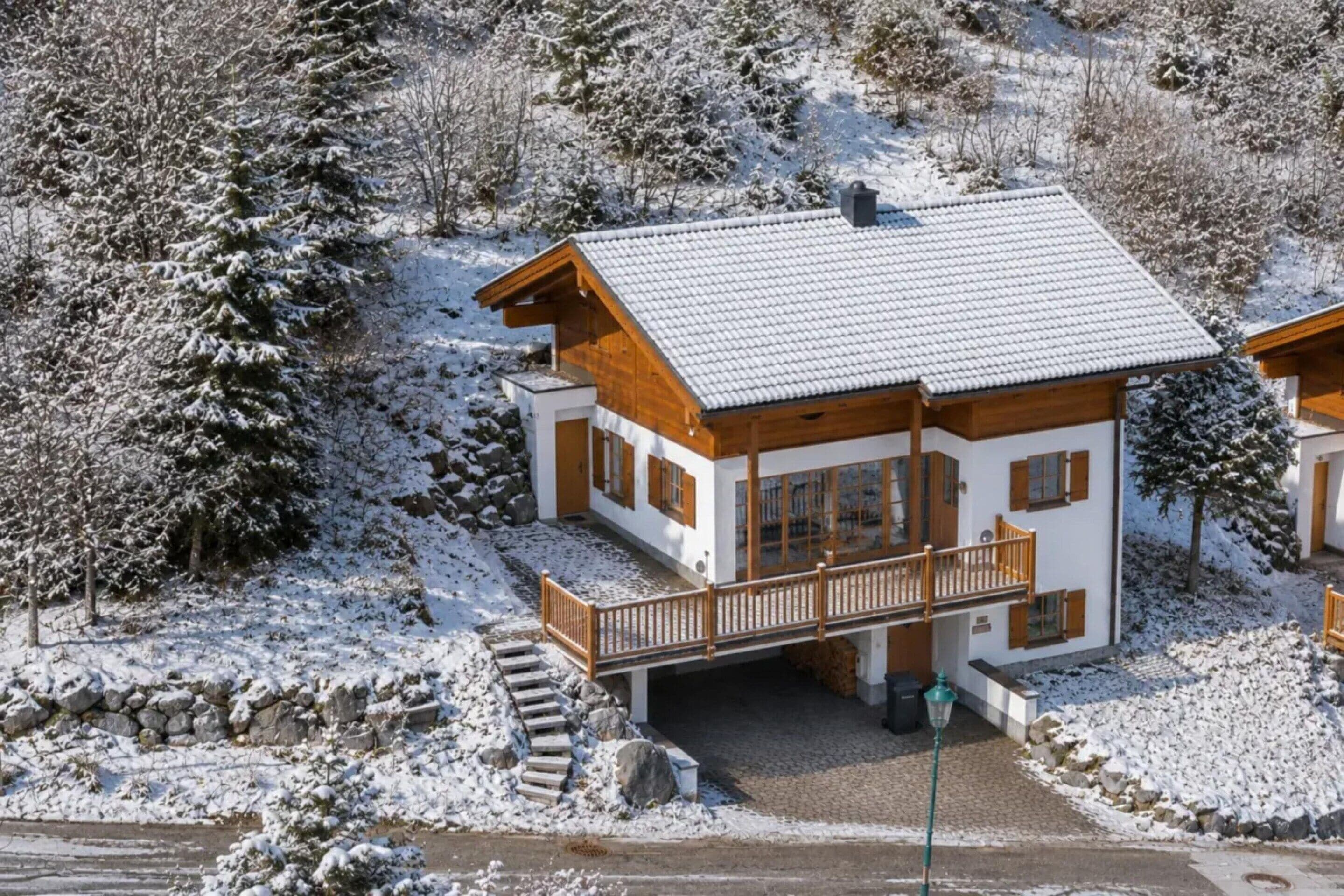 A two-story wooden chalet with a snow-covered roof, a balcony, and a stone staircase leading to the entrance.