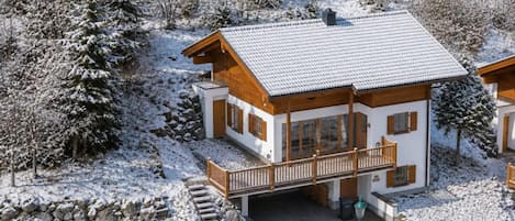 Un chalet en bois à deux étages, avec un toit enneigé, un balcon et un escalier en pierre menant à l’entrée.