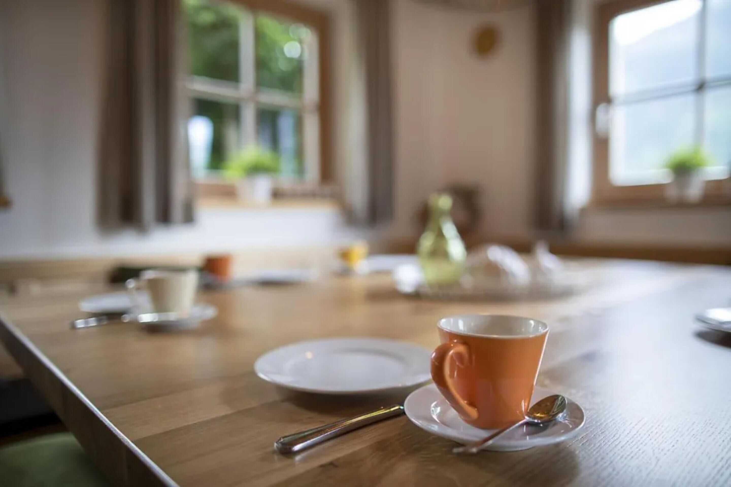 Una mesa de madera para comer con una taza y platillo, una cuchara y una tetera al fondo.