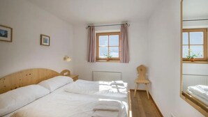 A bedroom with a wooden bed, a small chair, a window with curtains, and a mirror.