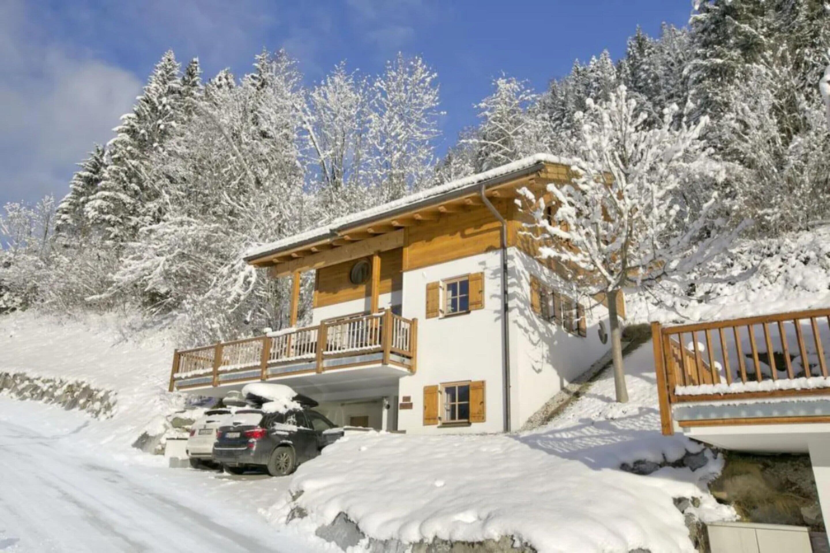 A two-story wooden chalet with a balcony, parked cars, and snow-covered trees.