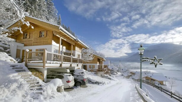 A wooden chalet with a balcony, parked cars, and a snow-covered landscape.