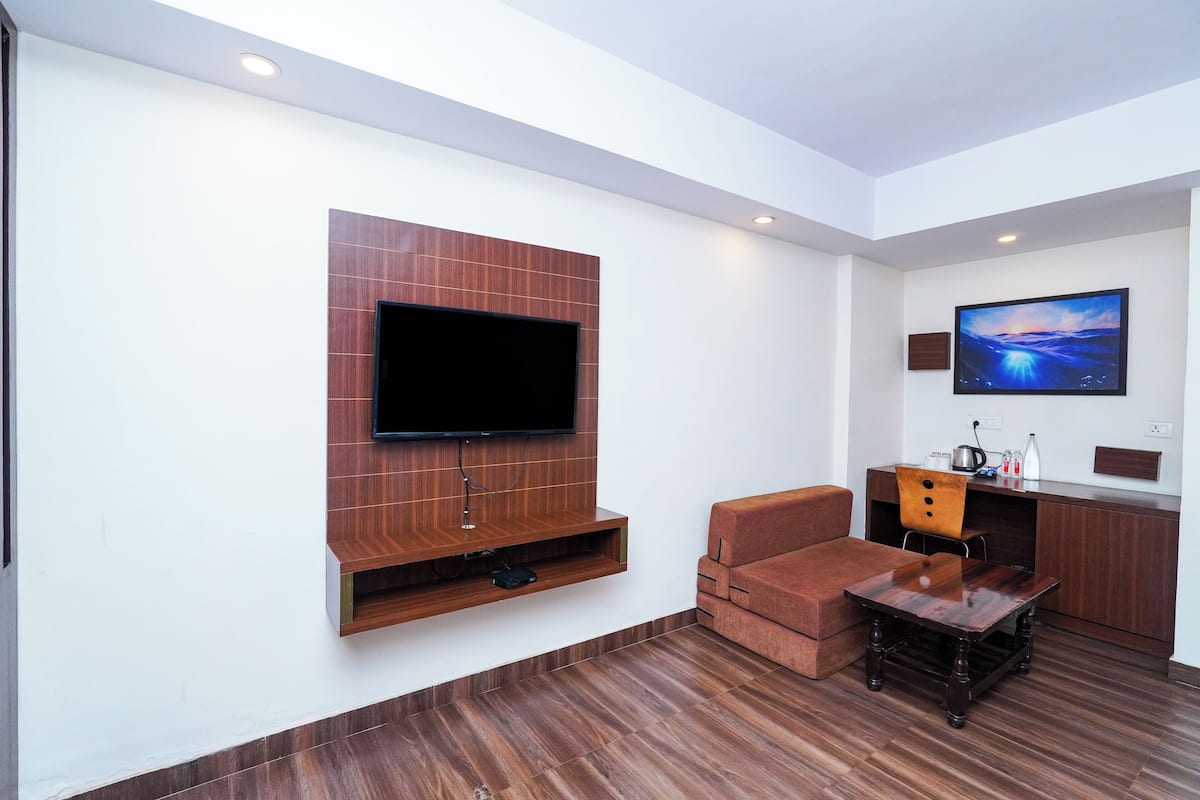 A modern living room with a flat-screen TV mounted on a wooden panel, a brown sofa, a wooden coffee table, and a small kitchen area with a microwave and kettle.