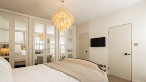 A bedroom with a large bed, a chandelier, a wardrobe with mirrored doors, and a television on the wall. - Welcome to your stylish retreat in the heart of Primrose Hill (London)