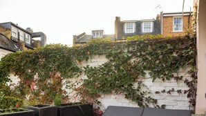 A rooftop terrace with a grey cushioned seating area, a white brick wall, and climbing plants. - Welcome to your stylish retreat in the heart of Primrose Hill (London)