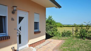 Una casa con puerta blanca, dos ventanas con contraventanas, base de ladrillo y un sendero pavimentado que lleva a un patio cubierto de césped con una cerca.