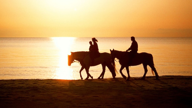 Dos personas cabalgando a caballo en una playa al atardecer.
