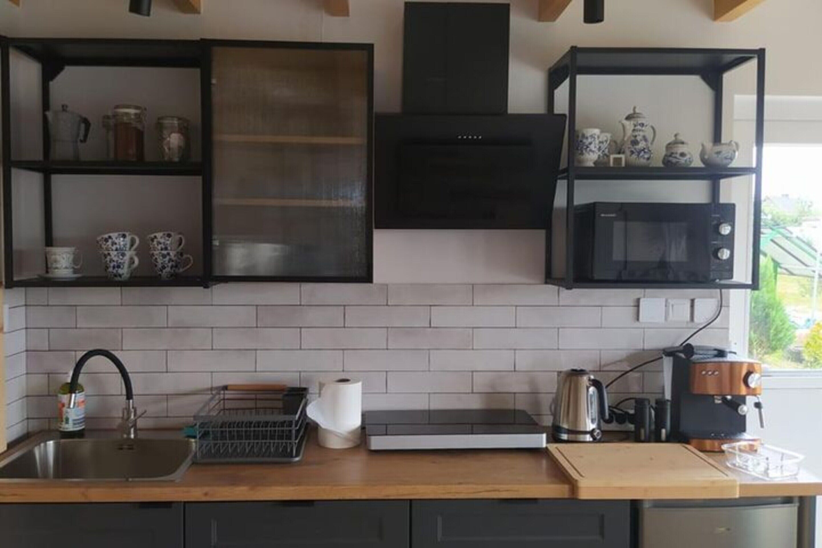 A modern kitchen with a built-in microwave, a coffee machine, and a kettle on the counter. There are shelves with kitchenware and a window providing natural light.
