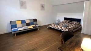 A room with a wooden floor, a sofa with a colorful cushion, a bed with a patterned cover, and a table with a lamp. - Apartment Saphir (5) 2/6 pers (Sedan)