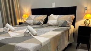 A neatly made bed with a patterned bedspread, two pillows, and rolled towels. There are two bedside lamps and a small table with a lamp. - Complete apartment with pool in Itajaí (Dom Bosco Itajaí)