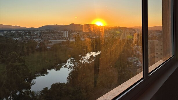 A view from a window overlooking a cityscape at sunset, with a river and hills in the foreground. - Complete apartment with pool in Itajaí (Dom Bosco Itajaí)
