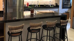 A modern kitchen with a stainless steel refrigerator, a built-in oven, and a gas stove. There are three bar stools with woven seats and black metal frames. The kitchen has pendant lighting and a stainless steel range hood. - Complete apartment with pool in Itajaí (Dom Bosco Itajaí)