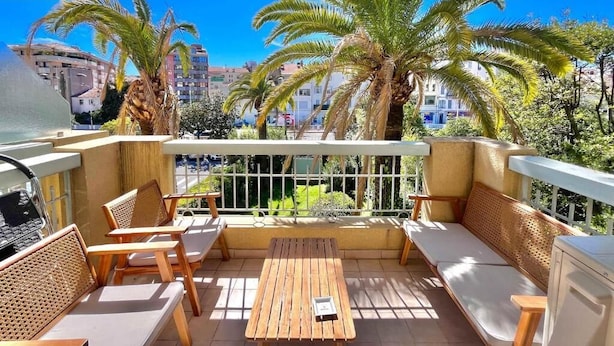 Un balcon doté de mobilier en bois, d’une table et offrant une vue sur des palmiers et des immeubles.