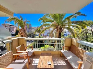 Un balcón con mobiliario de madera, una mesa y vistas a palmeras y edificios.