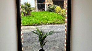 A corner room with a large window, a potted plant on a round wooden table, and a view of a stone building with a grassy yard. - Maison des H, by the lake (CANISY)