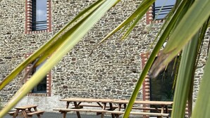 A stone building with a brick chimney, a window with a metal frame, and wooden picnic tables in front. - Maison des H, by the lake (CANISY)