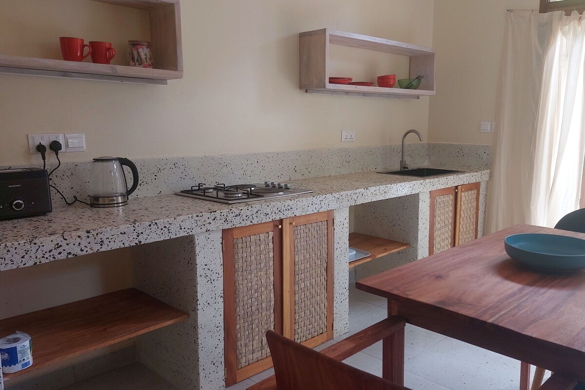 A compact kitchen with a gas stove, a kettle, and a sink. There are wooden cabinets with a woven design, a small shelf with red cups, and a table with a blue bowl.