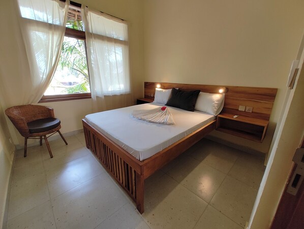 A single-bed room with a wooden headboard, a bedside table, a chair, and a window with sheer curtains. - Villa Bahati Zanzibar (Jambiani)