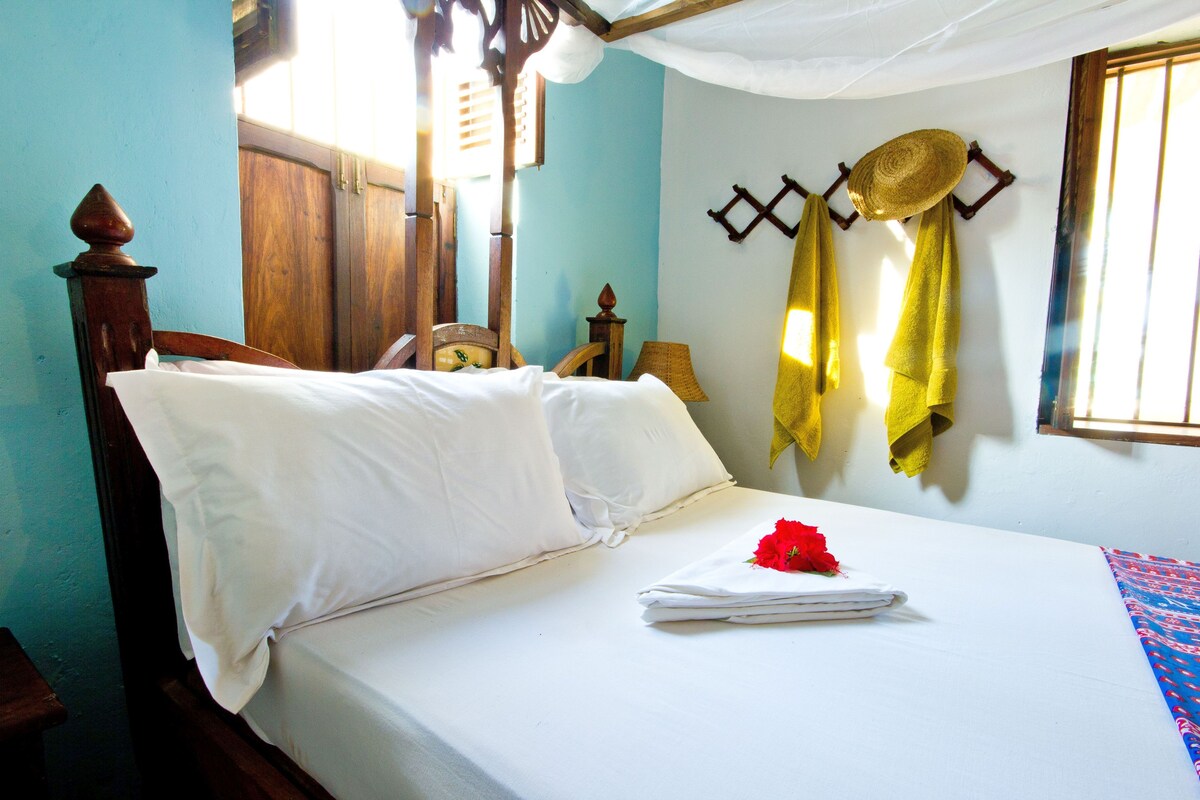 A neatly made bed with white linens, a red flower, and a yellow towel hanging on a wall hook.